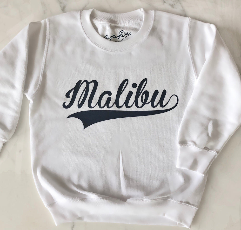 On The Rise Malibu Pullover Sweatshirt. A white sweatshirt with Malibu written in black cursive text across the chest. 