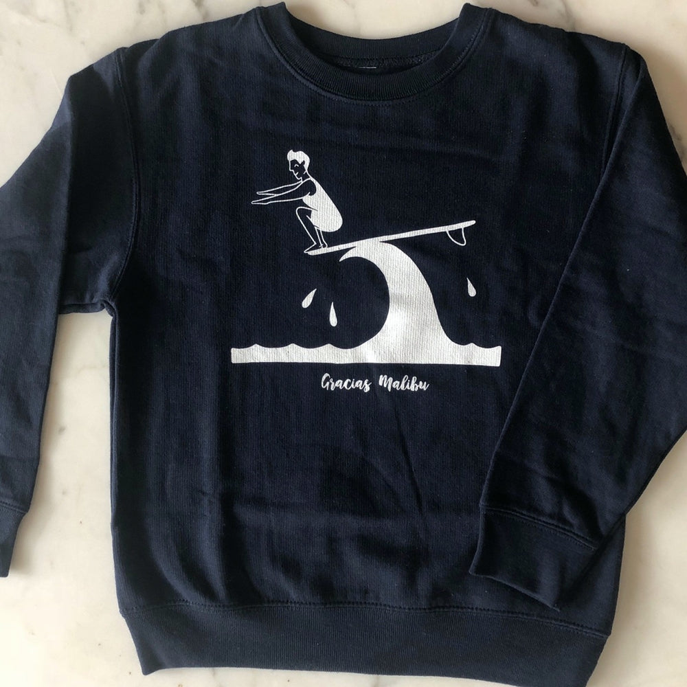 Kids size black crewneck sweater. There is an image of a woman in a bathing suit surfboarding on a wave. She is standing at the front point of the board crouched down with her arms out. The image is white in color. There is white text bellow the wave that says Gracias Malibu.