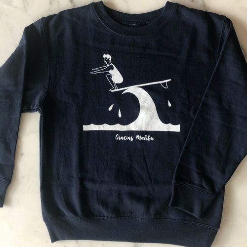 Kids size black crewneck sweater. There is an image of a woman in a bathing suit surfboarding on a wave. She is standing at the front point of the board crouched down with her arms out. The image is white in color. There is white text bellow the wave that says Gracias Malibu.