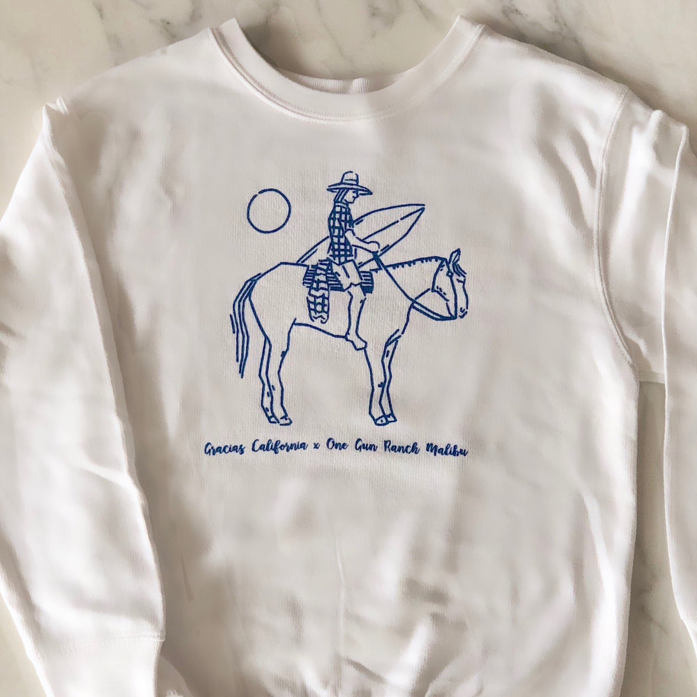 white sweatshirt with a blue outline drawing of a woman riding a horse and holding a surfboard. Underneath the text reads gracias california X one gun ranch malibu