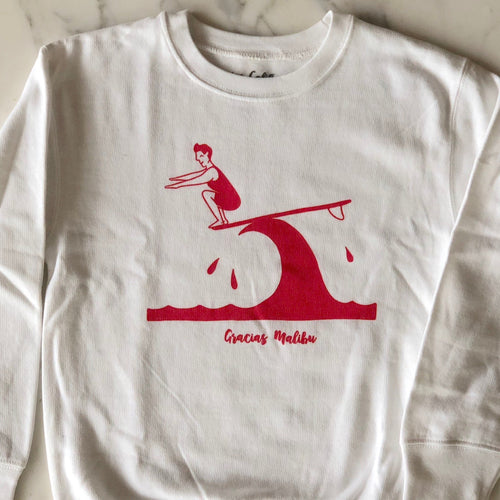 Gracias Malibu white sweatshirt with a red print design of a man surfing on a wave crouching down at the end of a surfboard. The text at the bottom reads gracias Malibu. 