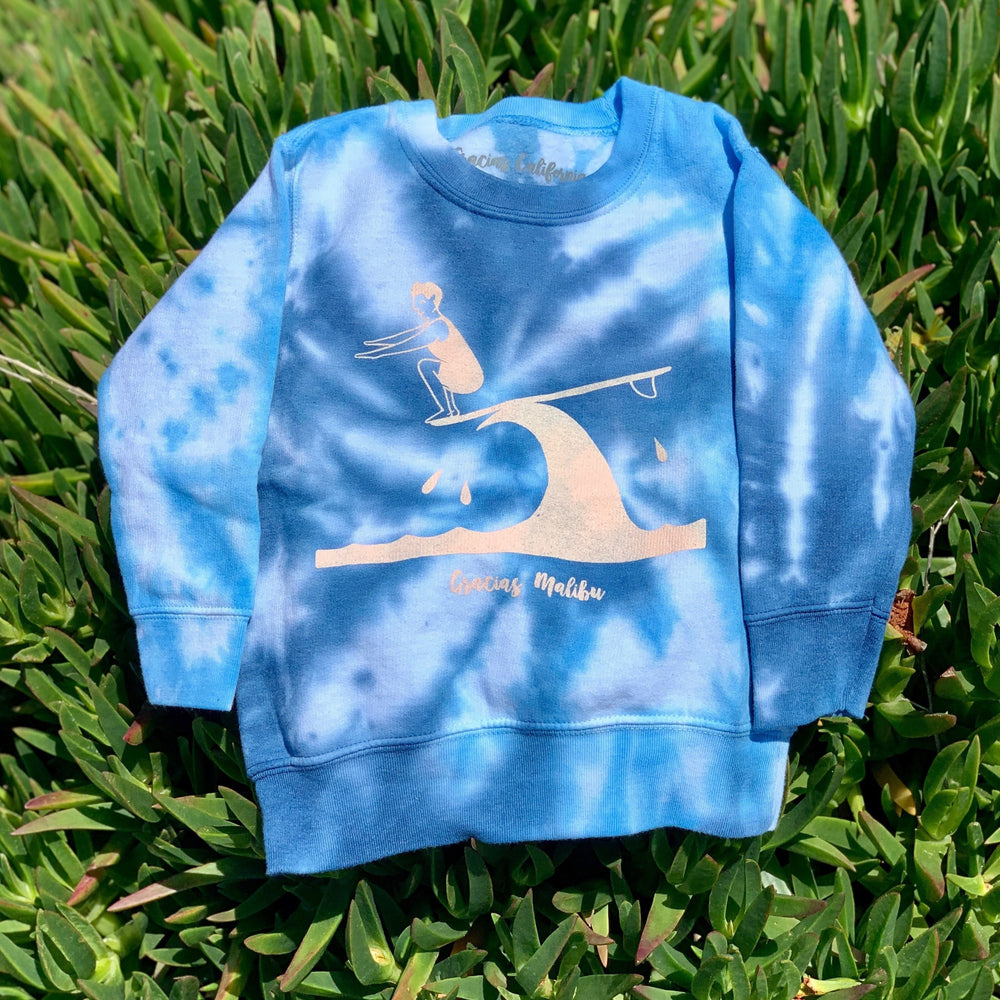 Kids Gracias Malibu blue and white tiedye sweatshirt with a white print design of a man surfing on a wave crouching down at the end of a surfboard. The text at the bottom reads gracias Malibu. 