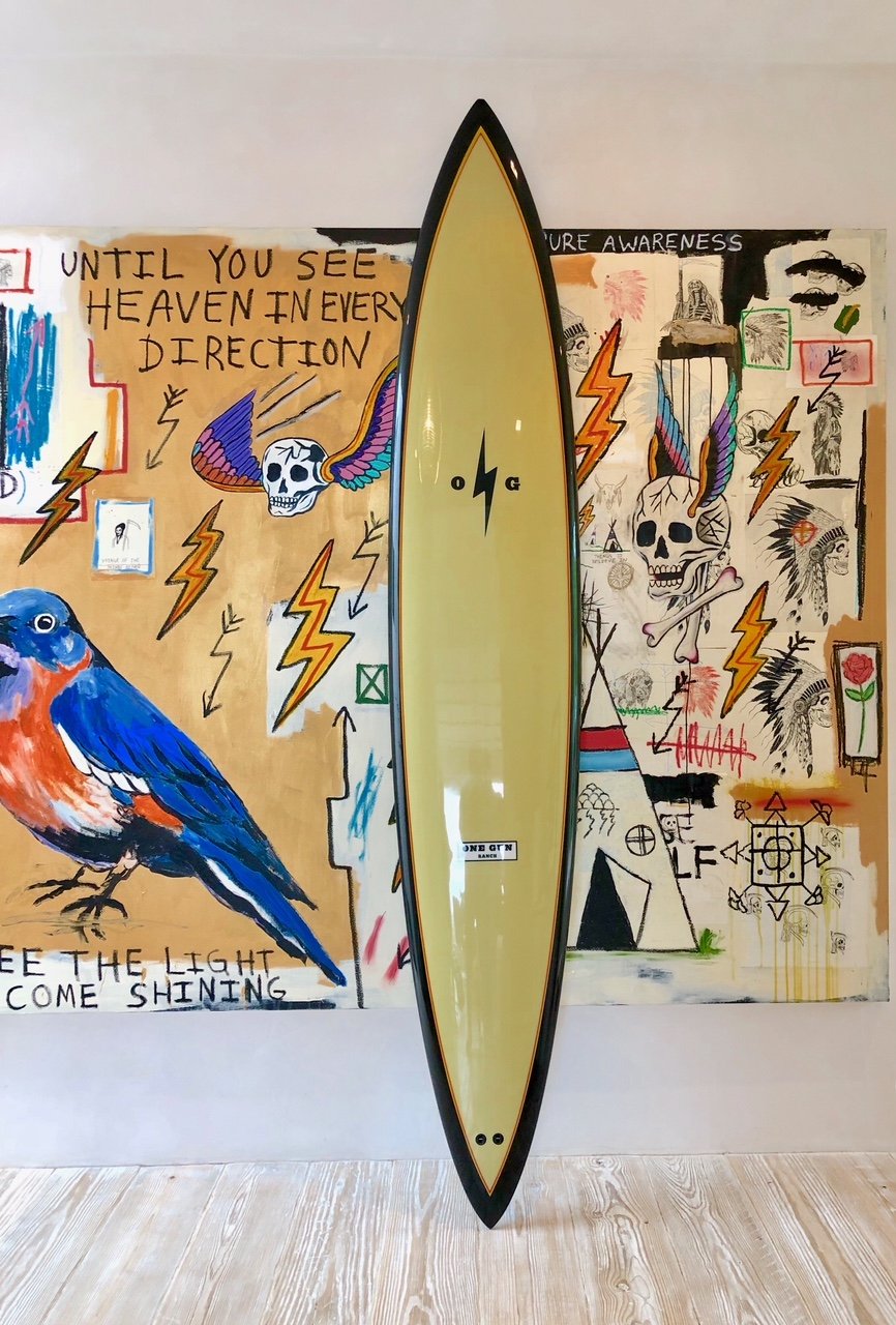 Light yellow long board surfboard. There is a black trim around the outside. There is a black lightning bold on the upper portion of the board towards the nose. There is a white and black logo on the back half of the board that says "One Gun Ranch. 