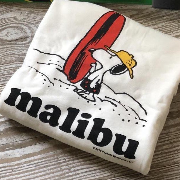 TSPTR Malibu Hot Dog T-Shirt. White shirt with a design of Snoopy holding what looks to be a surfboard, but is actually a hotdog. The word Malibu is written bellow him. 