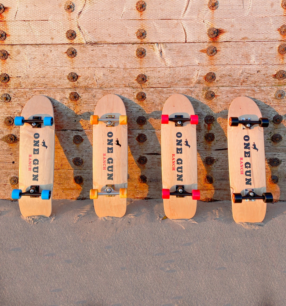 One Gun Ranch Buckshot x Arbor Skateboards. Brown wood boards with The One Gun Ranch logo on the bottom. Wheels are blue, orange, pink, and black. Bearings are silver and black .