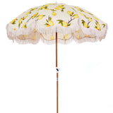 Business and pleasure vintage lemons beach umbrella. It has a canvas top with prints of lemons and leaves on it. There are white frills hanging down from the edges. The stand is a dark wood. 