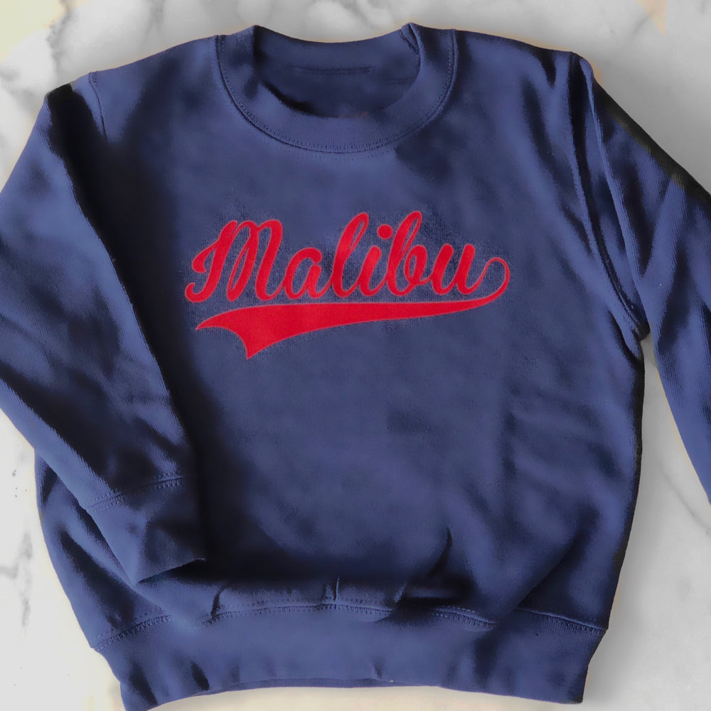 A blue sweatshirt with Malibu written on the chest in red cursive text. 