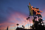 A photograph of a neon sign located on the streets of Los Angeles. There clouds are pink and the sky is blue. There are buildings on the bottom and palm trees in the distance. The sign has a neon yellow outlined surfboard, and red neon lettering saying "Safari Inn"