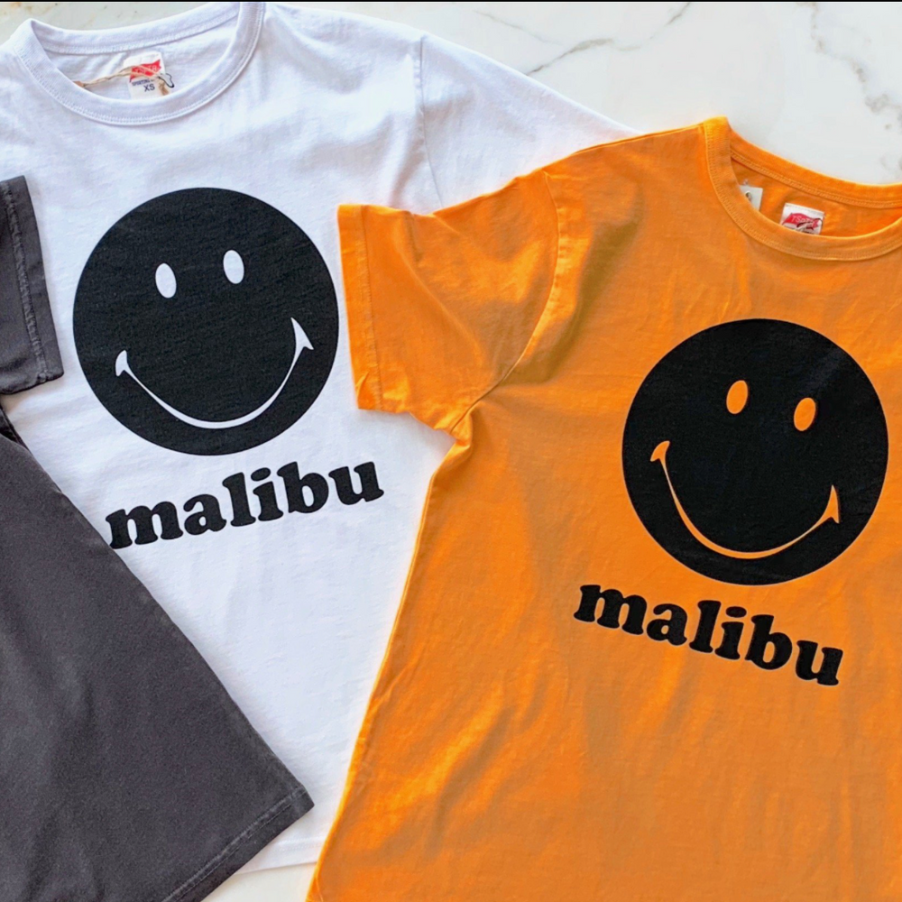 Grey, white and orange t-shirts with a black smily face in the center with the word Malibu underneath it. 