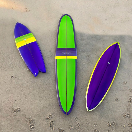 Three Album x One Gun Violet surfboards. The left is the smallest, the center is a long board, and the right is a medium board. They are all purple, green and yellow in design.