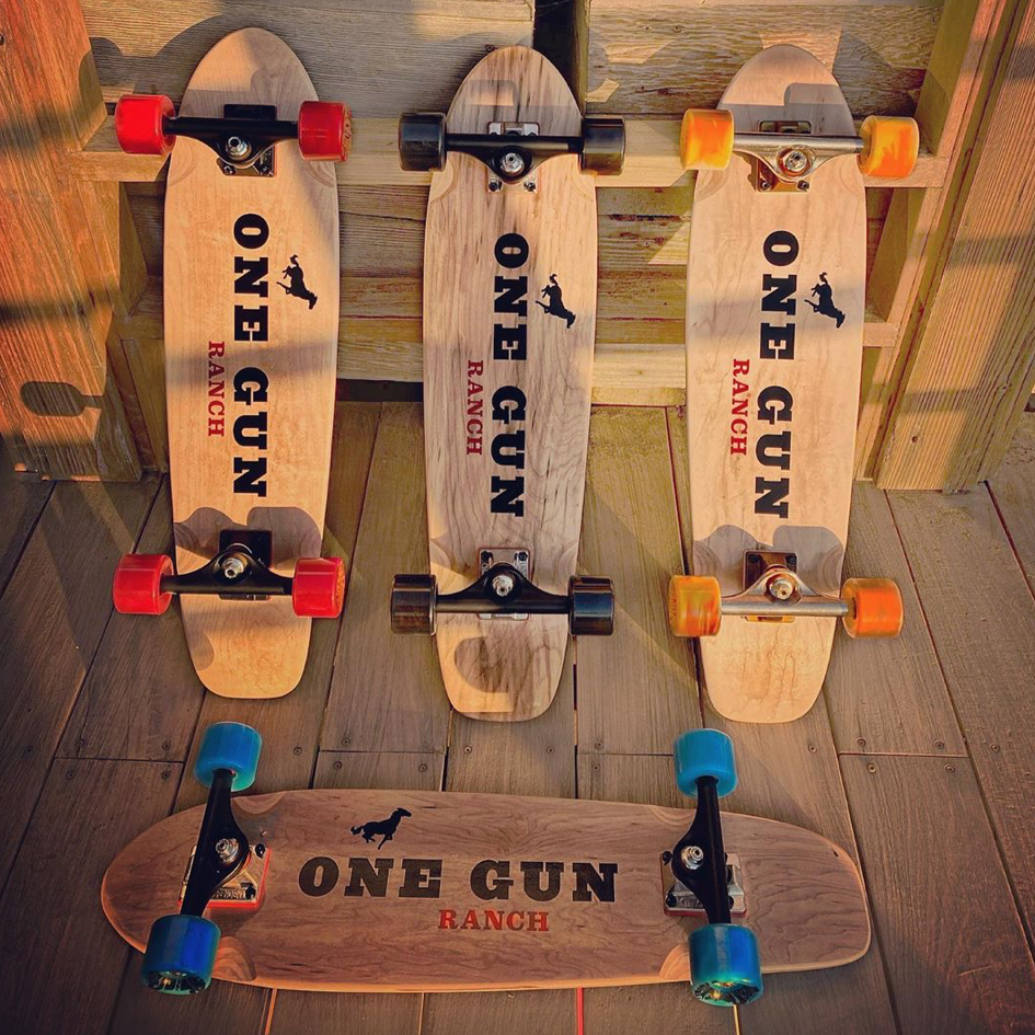 One Gun Ranch Buckshot x Arbor Skateboards. Brown wood boards with The One Gun Ranch logo on the bottom. Wheels are blue, orange, red, and black. Bearings are silver and black .