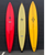 3 surfboards lined up. There's a red one on the left with a lightning bolt on the top center. The middle is a yellow board with black trim around the boarder. The right one is yellow with two lightning bolts and the logo One Gun in the middle. 