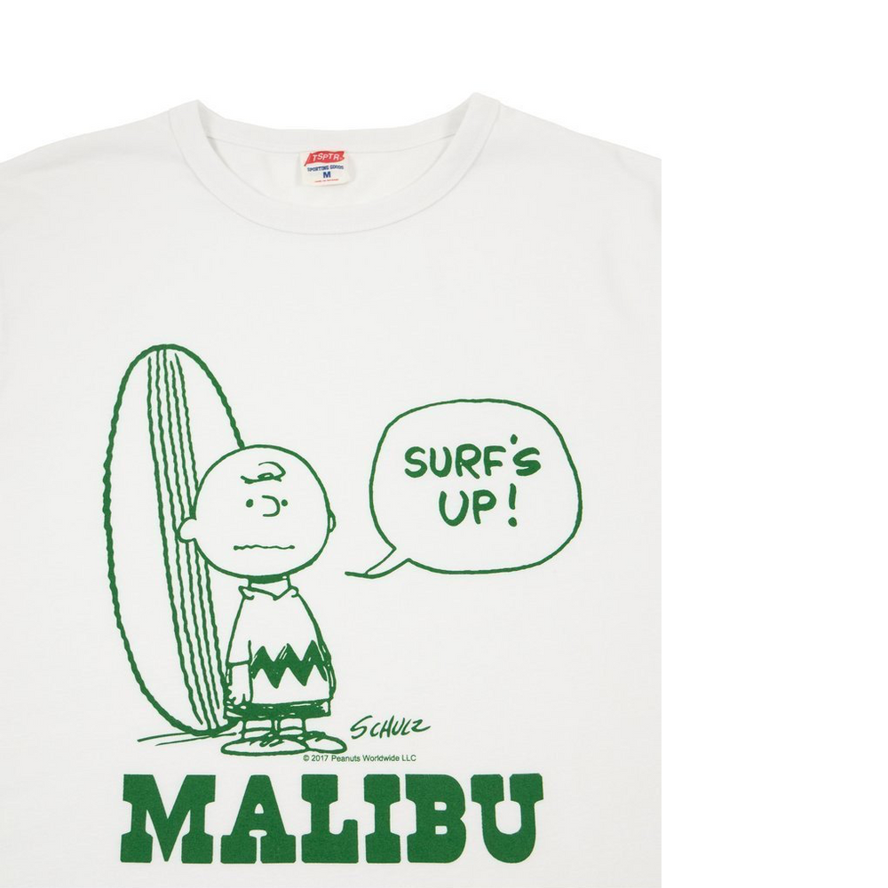 White t-shirt with a green design of Charlie Brown standing next to a surfboard with a word bubble of him saying Surf's Up!. Bellow him is the word Malibu