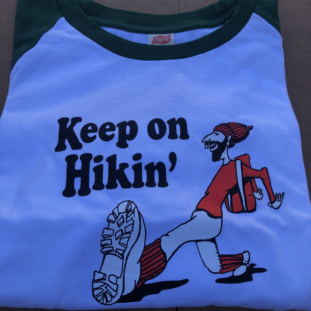 TSPTR Keep On Hikin Raglan T-Shirt. White shirt with green sleeves. On it is a design of a man wearing orange clothing. The words Keep On Hikin' in front of him. 